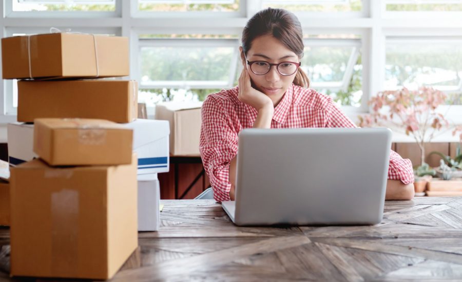 woman sitting at desk looking at laptop with large stack of parcels